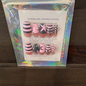 Puede incluir: Un juego de uñas postizas en un paquete de plástico transparente. Las uñas presentan varios diseños, incluyendo rayas negras y rosas, estrellas y lunares. El paquete incluye instrucciones de cuidado.
