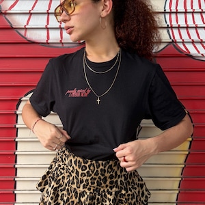 Puede incluir: Camiseta negra con la frase "proudly mixed by a Cuban mom" en letras rojas. La persona lleva un collar dorado con un colgante de cruz, pendientes de aro dorados y una falda con estampado de leopardo. También lleva gafas de sol tintadas de amarillo.