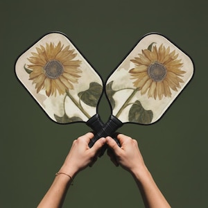 Könnte beinhalten: Zwei Pickleball-Schläger mit Sonnenblumen-Design. Die Schläger haben einen schwarzen Rand und schwarze Griffe. Die Sonnenblumenkunst zeigt gelbe Blütenblätter und eine braune Mitte. Der Hintergrund ist einfarbig dunkelgrün.
