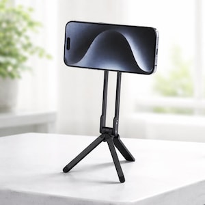 May include: A black smartphone is mounted on a black tripod stand. The phone displays a wavy, abstract image. The tripod has three legs and two vertical supports for the phone. The stand is on a white surface.