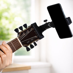 May include: Acoustic guitar headstock with a black smartphone holder attached. The guitar's headstock is dark brown with black tuning pegs. The smartphone is black and mounted at an angle, suggesting use for recording or viewing.