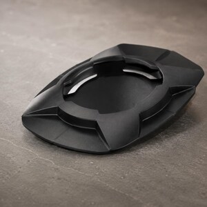 May include: A black ashtray with a geometric design. The ashtray has a central circular depression and several angled rests for cigarettes. The overall shape is a flattened oval with sharp, angular edges. The ashtray is made of a matte black material.