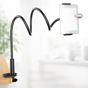 May include: A black, flexible tablet holder with a clamp base and a bendable arm. The holder secures a white tablet with black and pink clips. The clamp attaches to a wooden surface, offering a hands-free viewing experience.