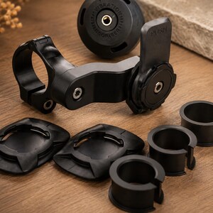 May include: Black motorcycle phone mount kit with various components. Includes a handlebar clamp, a phone holder with a "PUSH" label, vibration dampener, and several mounting adapters. The kit is designed to securely hold a phone while riding.