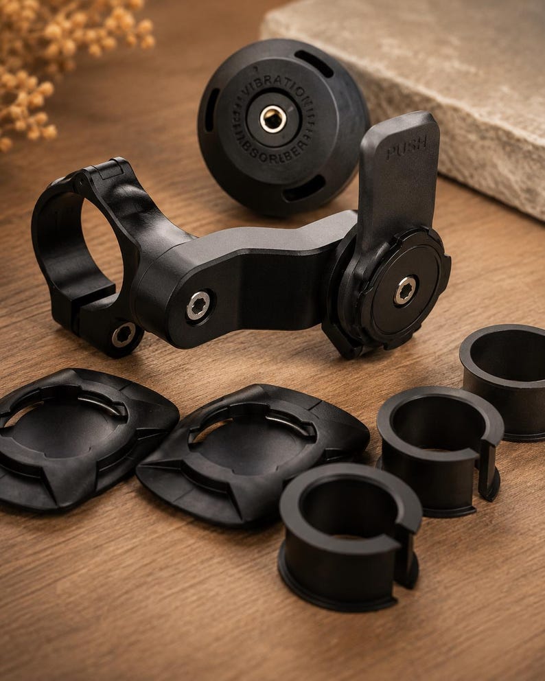 May include: Black motorcycle phone mount kit with various components. Includes a handlebar clamp, a phone holder with a "PUSH" label, vibration dampener, and several mounting adapters. The kit is designed to securely hold a phone while riding.