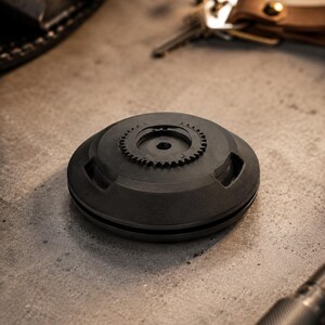 May include: A dark grey, circular object featuring a gear-shaped detail on its upper surface. The object has a matte finish and rests on a concrete surface. Keys and a leather keyring are in the background.