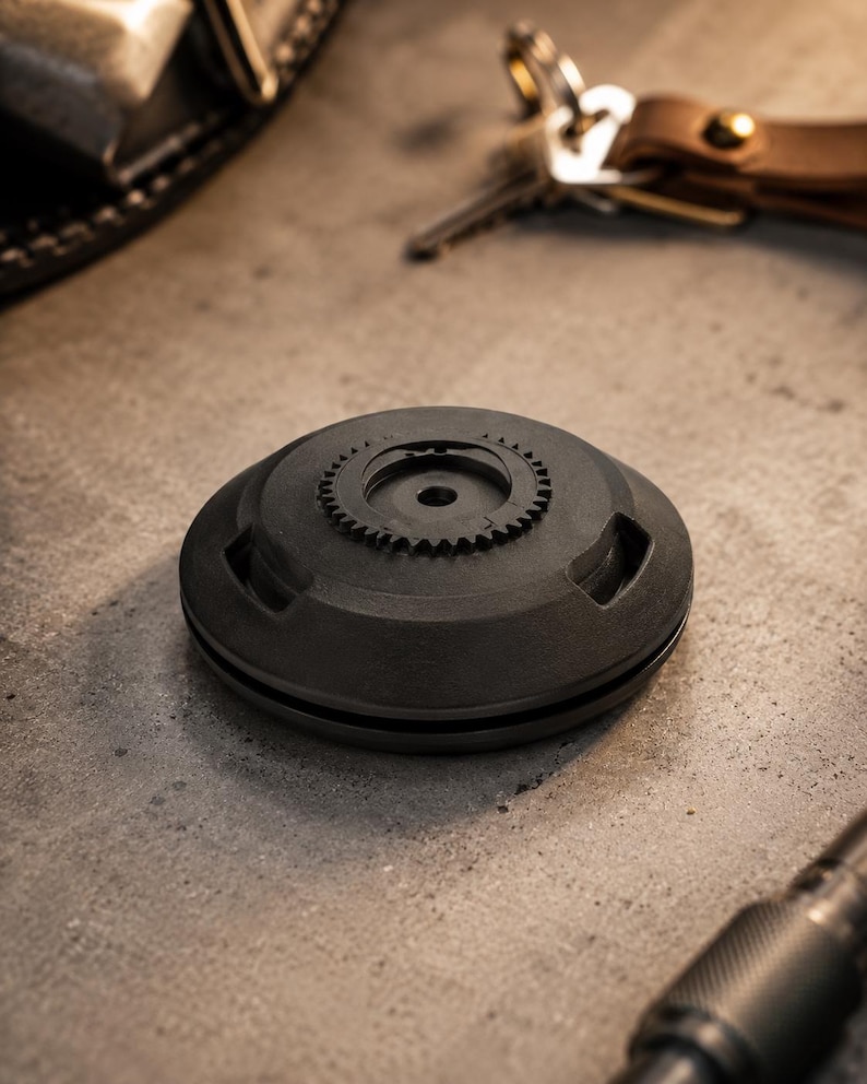 May include: A dark grey, circular object featuring a gear-shaped detail on its upper surface. The object has a matte finish and rests on a concrete surface. Keys and a leather keyring are in the background.
