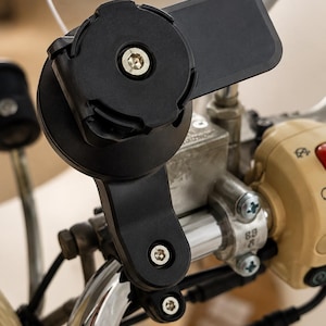 May include: A black motorcycle phone mount fixed to the handlebars. The mount features a circular base with a central screw and a rectangular extension. The handlebars are chrome, and the controls are beige.