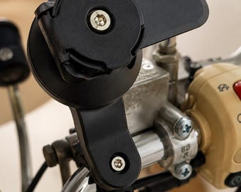 May include: A black motorcycle phone mount attached to the handlebars. The mount has a circular base with a central screw and a rectangular extension. The handlebars are chrome, and the controls are beige.