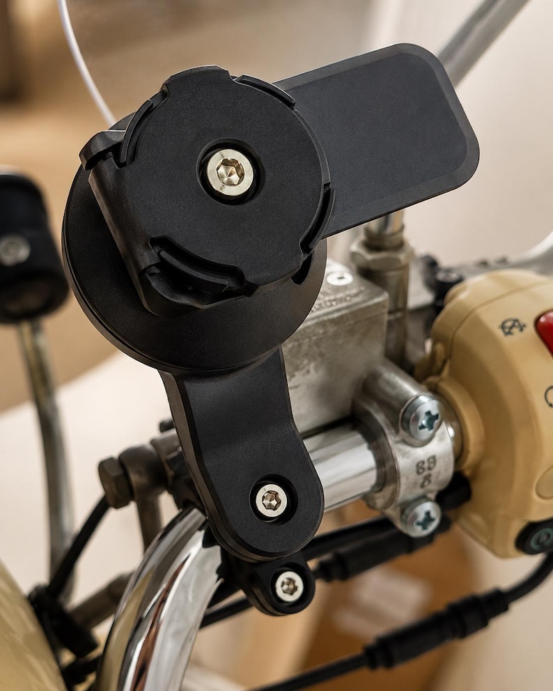 May include: A black motorcycle phone mount fixed to the handlebars. The mount features a circular base with a central screw and a rectangular extension. The handlebars are chrome, and the controls are beige.