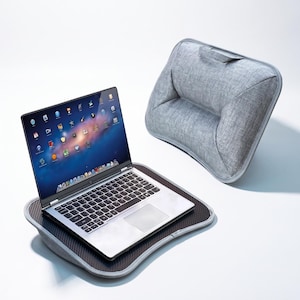 Puede incluir: Un portátil plateado con teclado negro y pantalla con iconos de aplicaciones sobre un soporte de fibra de carbono negra. El soporte incluye una base de cojín de tela gris con asa y un respaldo de tela gris a juego para mayor comodidad.