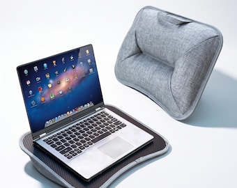 May include: A silver laptop with a black keyboard and screen displaying various application icons sits on a black carbon fiber textured lap desk. The lap desk features a gray fabric cushion base with a handle and a matching gray fabric back support.
