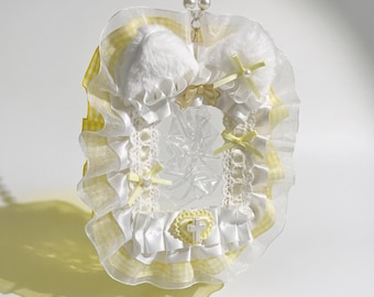 May include: A rectangular photo frame featuring a white faux fur top with cat ears, yellow and white gingham ruffles, white lace trim, and small yellow ribbon bows. A yellow heart-shaped charm with a white cross sits at the bottom center.