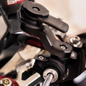 May include: Black motorcycle phone mount with several articulating joints and a circular mounting plate. The mount is attached to the motorcycle's handlebars and has a sleek, modern design. The SP logo is visible in the background.