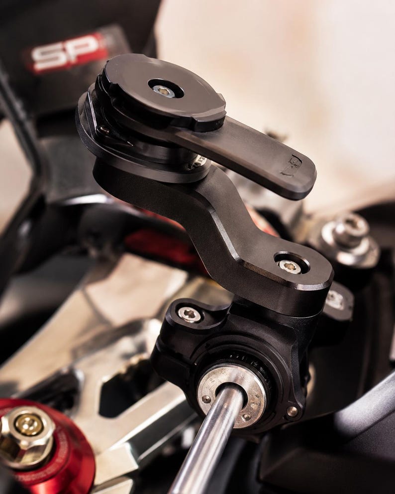 May include: Black motorcycle phone mount with several articulating joints and a circular mounting plate. The mount is attached to the motorcycle's handlebars and has a sleek, modern design. The SP logo is visible in the background.