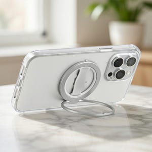 May include: A clear phone case with a silver ring holder attached to the back. The phone is propped up on a table. The ring holder can be used as a stand. The phone has three camera lenses.