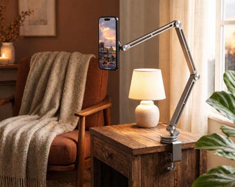 May include: A silver adjustable phone holder with a clamp base is attached to a wooden nightstand. A smartphone displays a cityscape. A small lamp with a beige shade sits on the nightstand. A brown armchair and a framed picture are in the background.