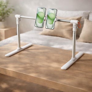 May include: A dual-phone stand with a white base and silver adjustable arms. Two smartphones are mounted on the stand, displaying green screens. The stand is on a wooden surface, with a bed and pillows in the background.