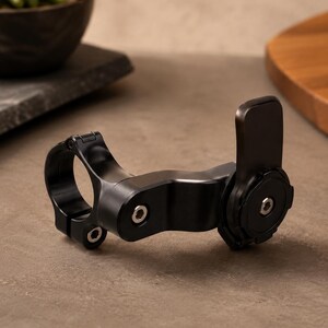 May include: A black metal bicycle handlebar mount with a circular clamp and a pivoting arm. The mount has a matte finish and is designed to hold a device. The mount is shown on a neutral background.