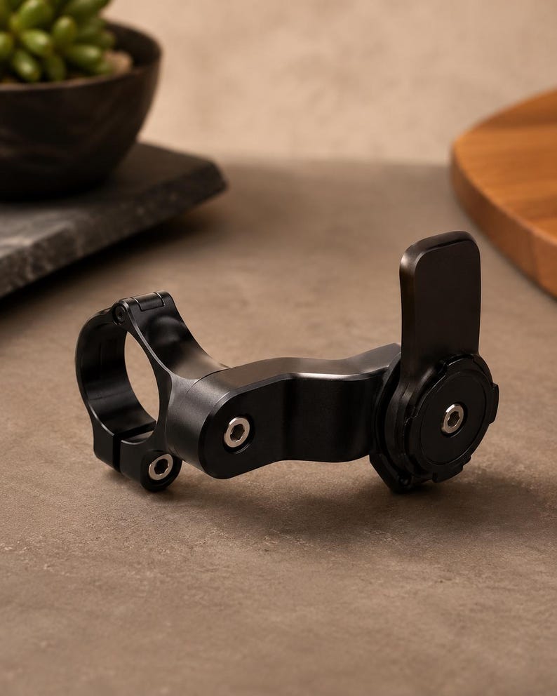 May include: A black metal bicycle handlebar mount with a circular clamp and a pivoting arm. The mount has a matte finish and is designed to hold a device. The mount is shown on a neutral background.