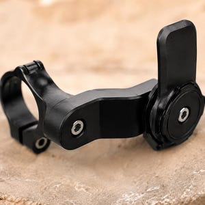 May include: A black bicycle handlebar mount with a circular clamp and a pivoting arm. The mount has a rectangular locking mechanism and several screw details. The product is designed to attach to a bicycle handlebar.