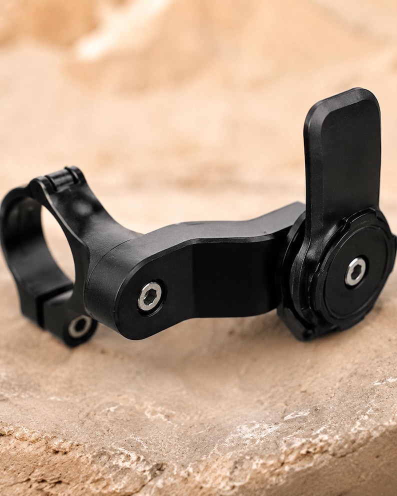 May include: A black bicycle handlebar mount with a circular clamp and a pivoting arm. The mount has a rectangular locking mechanism and several screw details. The product is designed to attach to a bicycle handlebar.