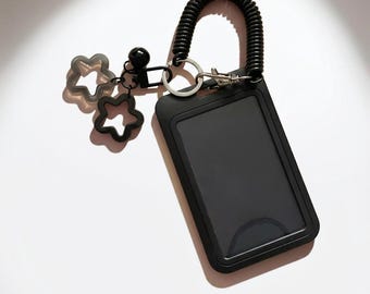 May include: A black rectangular ID badge holder with a clear window, attached to a black coiled wrist strap. The accessory features a silver metal ring, a black bead, and two small black star-shaped charms hanging from the side of the holder.