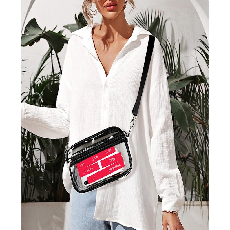 May include: A clear, rectangular crossbody bag with a black strap and trim. The bag contains red cards with white text. The bag is worn over the shoulder. The background is a white wall with green plants.