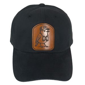May include: A black baseball cap featuring a tan leather patch on the front. The patch displays an engraved illustration of a bird wearing a cap that says BIRD NERD and carrying binoculars around its neck. The cap has a curved brim and stitched eyelets.