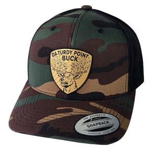 May include: Camouflage pattern trucker hat with a black mesh back. The front features a leather patch with the text "DA TURDY POINT BUCK" above a buck illustration. The hat has a curved brim and a snapback closure.