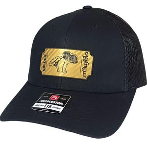 May include: Black trucker hat with a mesh back and a curved brim. The front features a gold patch with the words "GOLDEN CHANTERELLE" and illustrations of mushrooms. The hat also has a small white tag with the brand name "Richardson".