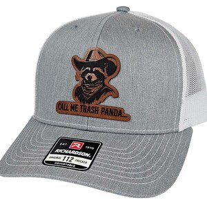 May include: Gray trucker hat with a white mesh back. The hat features a leather patch with a raccoon wearing a cowboy hat and the text "CALL ME TRASH PANDA...". The hat is adjustable.