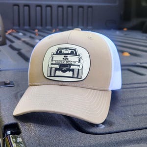 May include: Tan and white trucker hat with a curved brim. The hat features a patch with a black and white illustration of a truck and the words "SUPER BOOTY". The hat is sitting on a black surface.