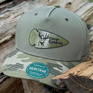 May include: An olive green baseball cap with a camouflage pattern on the brim. The front features an arrowhead-shaped patch with a deer illustration and the text Camp Coldren. A circular sticker on the brim reads Legacy Heritage.