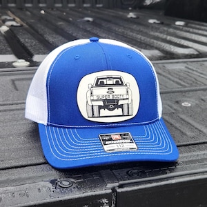 May include: A blue and white trucker hat with a patch featuring a truck graphic and the words "SUPER BOOTY". The hat has a curved brim and a mesh back. The Richardson 112 adjustable hat is visible.