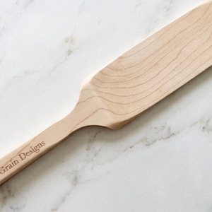 May include: A light-colored wooden kitchen spatula with a flat, rectangular head and a long handle. The handle is engraved with the text Bull & Grain Designs. The wood features a visible natural grain pattern throughout the entire utensil.