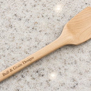 May include: A light brown wooden kitchen spatula with a flat, angled head and a long handle. The handle features the text Bull & Grain Designs engraved in a dark, clear font. The wood grain is visible throughout the surface of the utensil.
