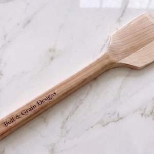 May include: A light-colored wooden spatula with a flat, angled head and a long handle. The handle features the engraved text Bull & Grain Designs and a small circular hole at the end. The wood grain is visible throughout the entire kitchen utensil.