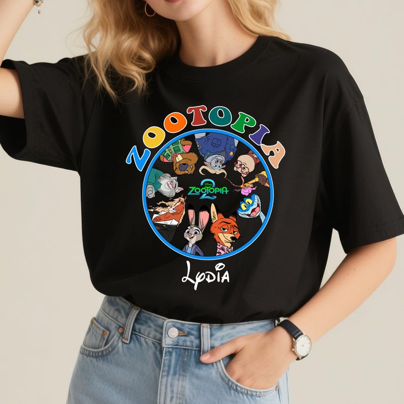 May include: Black t-shirt featuring a colorful circular graphic with characters from the animated film "Zootopia." The graphic includes the word "Zootopia" in a blue circle, with the text "Zootopia 2" and the name "Lydia" below.