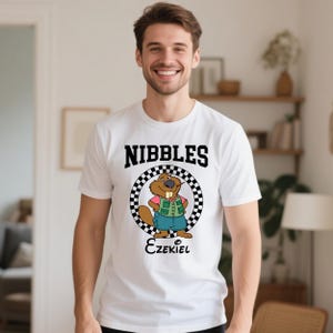 Puede incluir: Camiseta blanca con la palabra "NIBBLES" en negro sobre un personaje de castor de dibujos animados. El castor lleva una camisa rosa, un chaleco verde y pantalones cortos azules. El nombre "Ezekiel" est&aacute; escrito debajo.