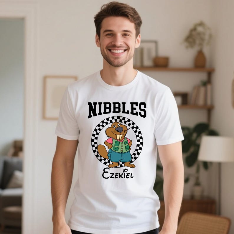 Puede incluir: Camiseta blanca con la palabra "NIBBLES" en negro sobre un personaje de castor de dibujos animados. El castor lleva una camisa rosa, un chaleco verde y pantalones cortos azules. El nombre "Ezekiel" est&aacute; escrito debajo.