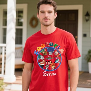 May include: A red t-shirt featuring a colorful circular graphic with characters from the movie "Zootopia". The word "Zootopia" is written in a circular pattern around the characters. The name "Steven" is printed in white script below the graphic.