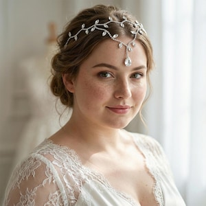 May include: A silver-toned bridal headpiece with a delicate leaf and teardrop design. The headpiece is adorned with clear crystals and is worn on the forehead. The woman in the image is wearing a white lace top.