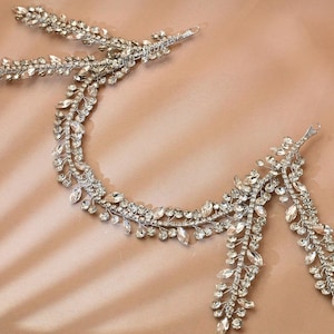 May include: A silver-toned bridal headpiece with a delicate leaf design. The headpiece is adorned with clear, sparkling crystals and small, round embellishments. The design includes two separate pieces, connected by a series of crystal-studded leaves.
