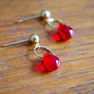 May include: A pair of gold-colored earrings with red teardrop-shaped pendants. The earrings have a post and ball design, with a small hoop connecting to the red glass pendant. The earrings are displayed on a wooden surface.