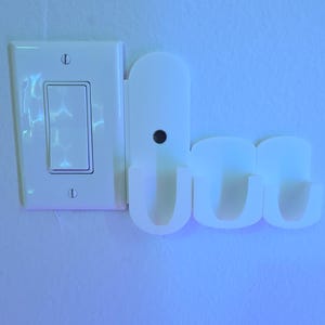 Puede incluir: Un interruptor de luz blanco y un soporte de plástico blanco con tres ranuras en forma de U están montados en una pared azul claro. El soporte está diseñado para sujetar objetos pequeños.
