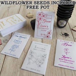 May include: A selection of wildflower seed packets, each personalized for a 30th birthday, with floral designs. The image also includes a stack of white packets with gold text, a small flag, and a set of black plant pots.