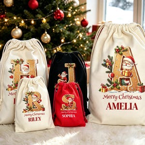 May include: A collection of five drawstring gift bags in cream, black, and red fabric. Each bag features a large gingerbread-style letter with a Santa Claus illustration, the text Merry Christmas, and a personalised name printed in red or white.