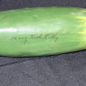 May include: A green, elongated object with a yellow-tinged end. The object has the handwritten text "mary Keck Kelly" in black ink. The object is set against a dark background.