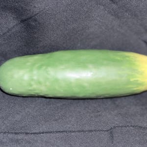May include: A green ceramic cucumber with a yellow tip rests on a black surface. The cucumber is smooth and elongated, with a gradient of color from green to yellow. This decorative item could be used as a kitchen or home decor accent.